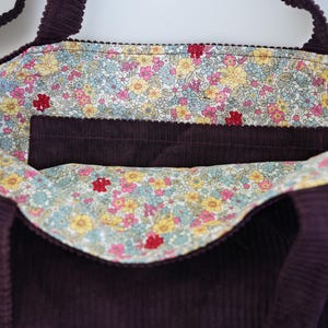 Op de afbeelding: Een dieppaarse corduroy tote bag met een bloemenvoering. De binnenkant heeft een zak en een kleurrijke bloemenstof met roze, gele en blauwe bloemen. De tas heeft twee handvatten.