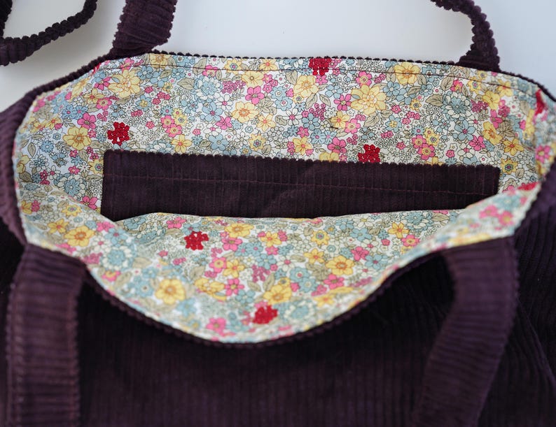 Op de afbeelding: Een dieppaarse corduroy tote bag met een bloemenvoering. De binnenkant heeft een zak en een kleurrijke bloemenstof met roze, gele en blauwe bloemen. De tas heeft twee handvatten.
