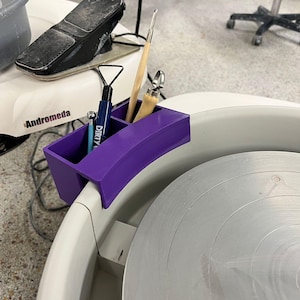 May include: A purple pottery tool holder attached to a white pottery wheel. The holder contains several tools, including a blue tool with the word "DIRTY" printed on it. A black foot pedal is visible in the background.