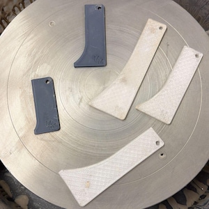 May include: Four pottery rib tools arranged on a circular surface. Two tools are dark gray with the letters MM embossed, and two are off-white with a textured, cross-hatched surface. Each tool features a curved edge and a small circular hole.