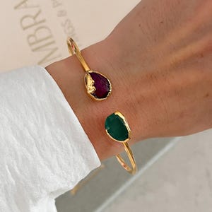 Op de afbeelding: Een goudkleurige open armband met twee asymmetrische, ruw geslepen edelstenen. De ene steen is diep verzadigd paars, terwijl de andere levendig groen is. De metalen band heeft een gladde, gepolijste afwerking en de stenen zitten in gestructureerde, organisch ogende gouden zettingen.