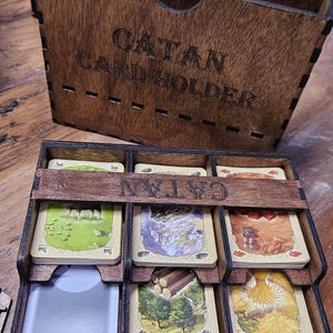 May include: A dark brown wooden card holder for the board game Catan. The box features the text CATAN CARD HOLDER engraved on the front. A separate tray with three columns holds various game cards, with the word CATAN engraved on the crossbar.