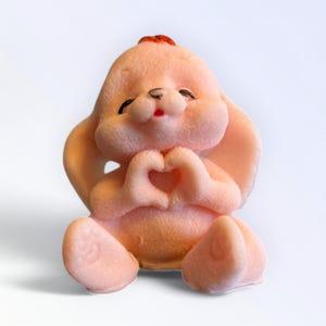 May include: A light pink bunny figurine with a heart-shaped gesture formed by its hands. The bunny has large ears, a red tuft of hair, and a smiling face with closed eyes. The figurine is sitting down.