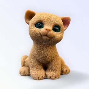 May include: A tan-colored cat figurine with large, expressive green eyes. The cat is sitting upright with its paws in front, and its ears are perked up. The figurine has a textured, fur-like appearance, and is set against a white background.