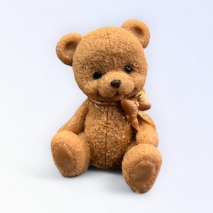May include: A brown teddy bear figurine with a textured surface, small black eyes, and a golden bow. The bear is sitting with its legs extended and arms at its sides. The bear is a classic toy.