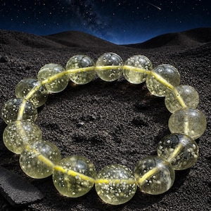 Puede incluir: Una pulsera circular compuesta por cuentas esféricas translúcidas de color amarillo pálido. Cada cuenta presenta una textura interna distintiva con pequeñas inclusiones blancas y moteadas en todo el material. Las cuentas están ensartadas en un cordón amarillo visible.
