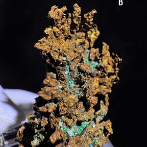 Puede incluir: Primer plano de un esp&eacute;cimen mineral de cobre con una superficie rugosa y texturizada. El mineral muestra una mezcla de tonos dorados y marrones met&aacute;licos, con parches de verde turquesa vibrante. El fondo es de color oscuro.