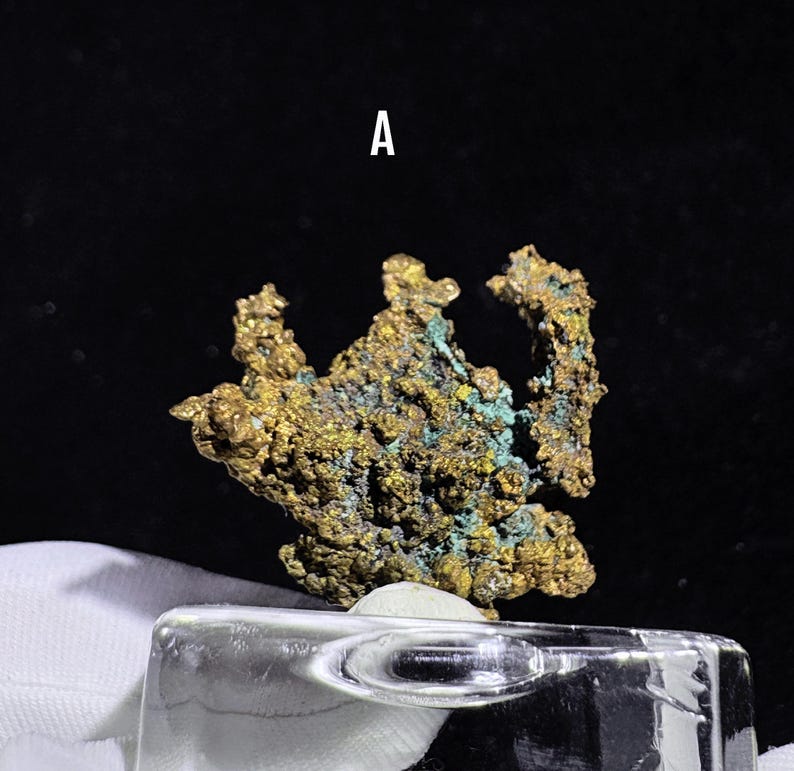 Puede incluir: Primer plano de un esp&eacute;cimen mineral con una superficie rugosa y texturizada. El mineral muestra una mezcla de colores dorados y turquesas, con la letra "A" en blanco en la parte superior. El esp&eacute;cimen se encuentra sobre una superficie transparente similar al vidrio.