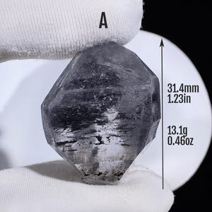 Peut inclure: Un cristal gris foncé, translucide et à multiples facettes étiqueté A. Le cristal mesure 31,4 mm de hauteur et pèse 13,1 g. Il présente des inclusions internes et une forme géométrique avec des bords distincts et des surfaces planes.