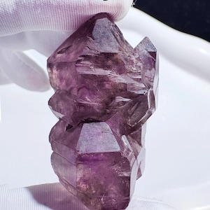 May include: A raw amethyst crystal cluster, displaying a deep purple colour with crystalline structures. The natural formation has a rough, textured surface and a faceted appearance. The letter "B" is visible in the upper portion of the image.
