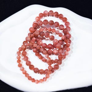 May include: Multiple strands of reddish-brown gemstone bead bracelets are displayed on a white, curved surface. The beads vary in size, creating a layered effect. The bracelets are likely made of sunstone or a similar material.