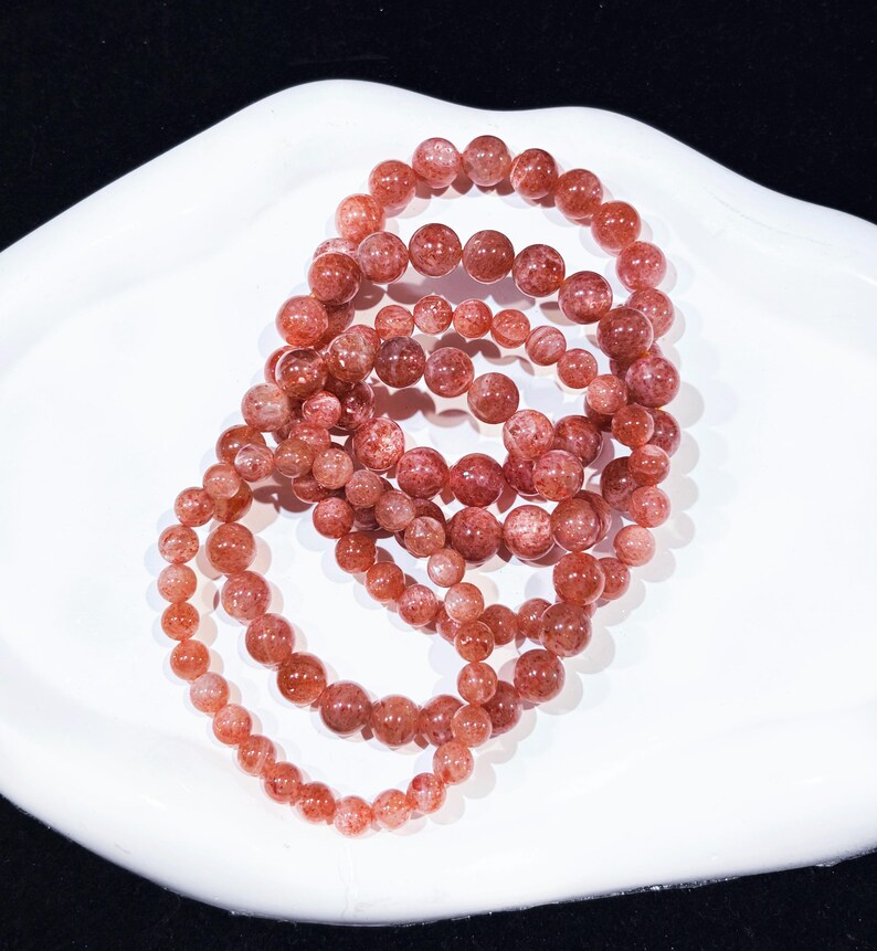 May include: Multiple strands of reddish-brown gemstone bead bracelets are displayed on a white, curved surface. The beads vary in size, creating a layered effect. The bracelets are likely made of sunstone or a similar material.