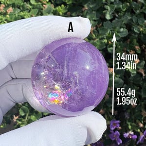 Peut inclure: Une sphère violette translucide étiquetée A, présentant des fractures internes et des reflets irisés. La sphère mesure 34 mm de diamètre et pèse 55,4 g, comme indiqué par le texte et le graphique en flèche à côté de l'objet.