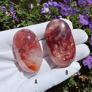 Peut inclure: Deux pierres de quartz ovales polies, rouges et transparentes, étiquetées A et B. Les pierres présentent des inclusions rouge foncé et des fractures internes au sein d'une structure cristalline translucide, posées sur un tissu blanc.