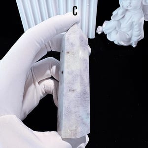 Puede incluir: Una torre de cristal morado claro y blanco con una parte superior puntiaguda. El cristal tiene forma rectangular y est&aacute; sostenido por una mano enguantada. La letra "C" es visible en la parte superior de la imagen.