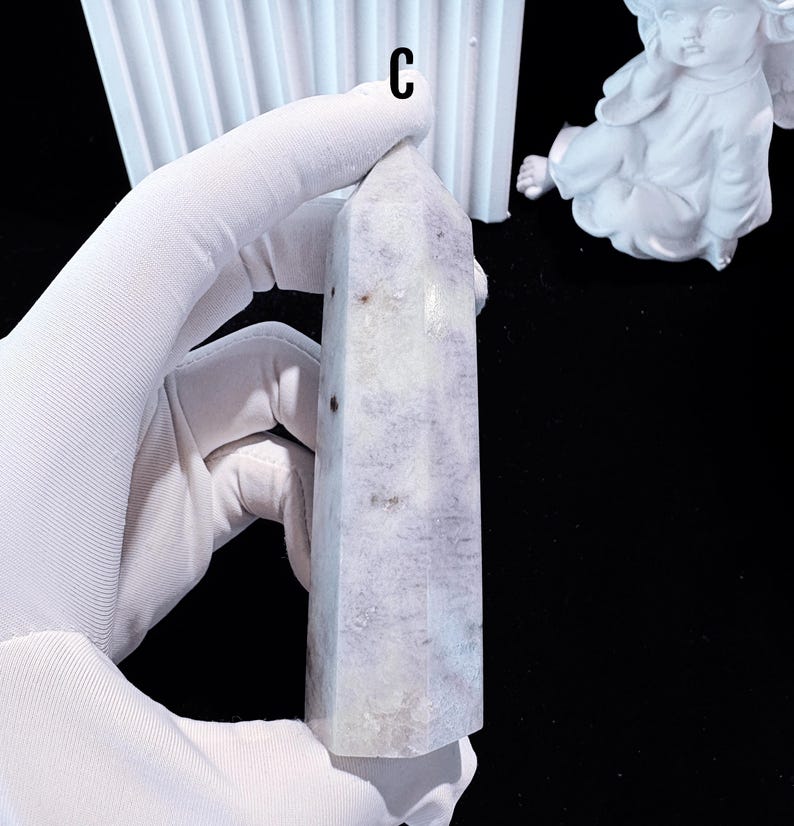 Puede incluir: Una torre de cristal morado claro y blanco con una parte superior puntiaguda. El cristal tiene forma rectangular y est&aacute; sostenido por una mano enguantada. La letra "C" es visible en la parte superior de la imagen.