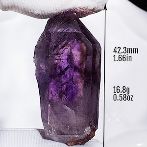 May include: A faceted, purple amethyst crystal is held up against a white background. The crystal is 42.3mm long and weighs 16.8g. The crystal has a deep purple color with lighter shades throughout. The length is also shown as 1.66in.