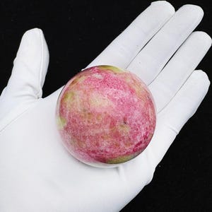 May include: A spherical gemstone, polished to a smooth finish, displaying a blend of pink and green colours. The stone is held in a white gloved hand against a black backdrop. The sphere has a marbled surface.