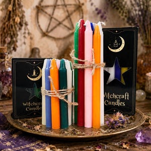 Può includere: Due fasci di candele affusolate multicolori legate con spago, posizionati davanti a due scatole rettangolari nere etichettate Witchcraft Candles con un design ritagliato a forma di mezzaluna e stella dorata, su un vassoio metallico rotondo.