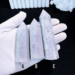 Puede incluir: Tres obeliscos de cristal morado claro con puntas. Los cristales se sostienen en una mano enguantada de blanco. Los obeliscos est&aacute;n etiquetados A, B y C. Los cristales tienen un aspecto veteado con sutiles variaciones de color.