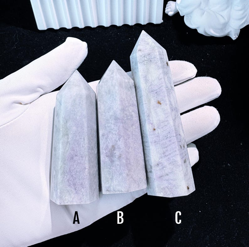 Puede incluir: Tres obeliscos de cristal morado claro con puntas. Los cristales se sostienen en una mano enguantada de blanco. Los obeliscos est&aacute;n etiquetados A, B y C. Los cristales tienen un aspecto veteado con sutiles variaciones de color.