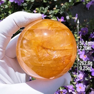 May include: A polished golden-orange sphere with internal fractures and inclusions. The sphere measures 57.4mm in diameter and weighs 262.6g. The surface is smooth and translucent, displaying varying shades of amber and honey tones throughout.