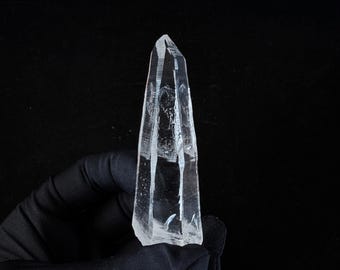 May include: A clear quartz crystal point, held against a black background. The crystal is transparent with visible internal structures and facets. The pointed end is sharply defined, and the overall shape is elongated and tapered.
