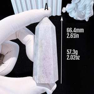 Puede incluir: Un obelisco de cristal morado claro, de 66,4 mm de altura y 57,3 g de peso. El obelisco tiene un dise&ntilde;o facetado y est&aacute; sostenido por una mano enguantada. El texto "A" es visible en la parte superior.