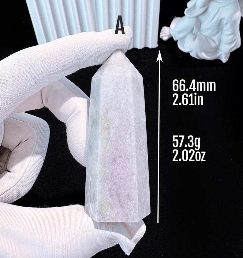 Puede incluir: Un obelisco de cristal morado claro, de 66,4 mm de altura y 57,3 g de peso. El obelisco tiene un dise&ntilde;o facetado y est&aacute; sostenido por una mano enguantada. El texto "A" es visible en la parte superior.