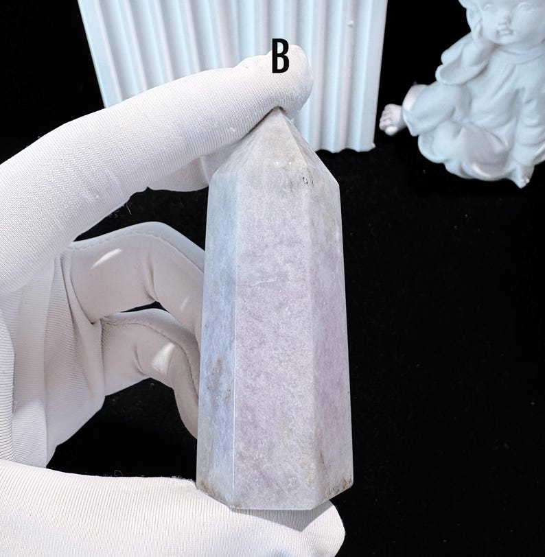 Puede incluir: Un obelisco de cristal morado claro con una base hexagonal y una parte superior puntiaguda. El cristal est&aacute; sostenido por una mano enguantada. El obelisco mide aproximadamente 10 cm de altura. El fondo incluye una pieza decorativa blanca y una superficie negra.