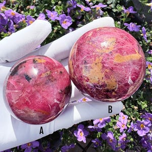 Puede incluir: Dos piedras esféricas pulidas con tonos magenta y rosa intenso con inclusiones naturales de depósitos minerales amarillos y negros. La esfera etiquetada como A es más pequeña que la esfera B, ambas con una superficie lisa y brillante.