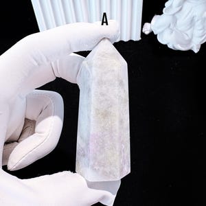 Puede incluir: Un obelisco de cristal rectangular de color claro con una parte superior puntiaguda. El cristal tiene una calidad sutil y transl&uacute;cida con toques de lavanda y gris. El obelisco est&aacute; sostenido por una mano enguantada, con un fondo negro.