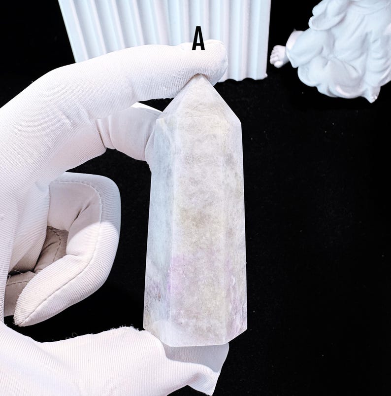 Puede incluir: Un obelisco de cristal rectangular de color claro con una parte superior puntiaguda. El cristal tiene una calidad sutil y transl&uacute;cida con toques de lavanda y gris. El obelisco est&aacute; sostenido por una mano enguantada, con un fondo negro.