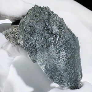 May include: A dark grey mineral specimen with an irregular shape and a rough, textured surface. The mineral is held in a white gloved hand, highlighting its natural form. The specimen has a crystalline structure and a metallic sheen.