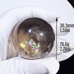 May include: A translucent, smoky brown quartz sphere featuring internal iridescent rainbow inclusions. The sphere measures 38.3 mm in diameter and weighs 78.6 g, as indicated by the text and arrow graphic visible next to the object.