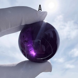 Puede incluir: Una piedra esférica pulida de color púrpura intenso marcada con la letra A. La superficie de la esfera es lisa y reflectante, mostrando inclusiones internas y refracción de la luz. El color violeta oscuro transiciona a tonos translúcidos.