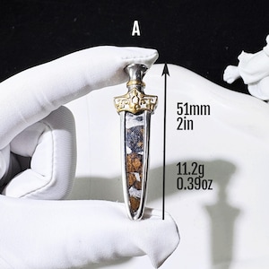 May include: A dagger-shaped pendant labelled A, featuring a silver frame with a gold-coloured crossguard and a textured, dark brown and silver inlay. The pendant measures 51mm in length and weighs 11.2g as indicated by the text.