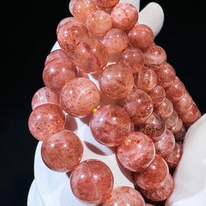 May include: A close-up of a bracelet made of round, reddish-orange beads. The beads have a speckled appearance and are strung together on a thin, orange cord. The bracelet is held against a white background.