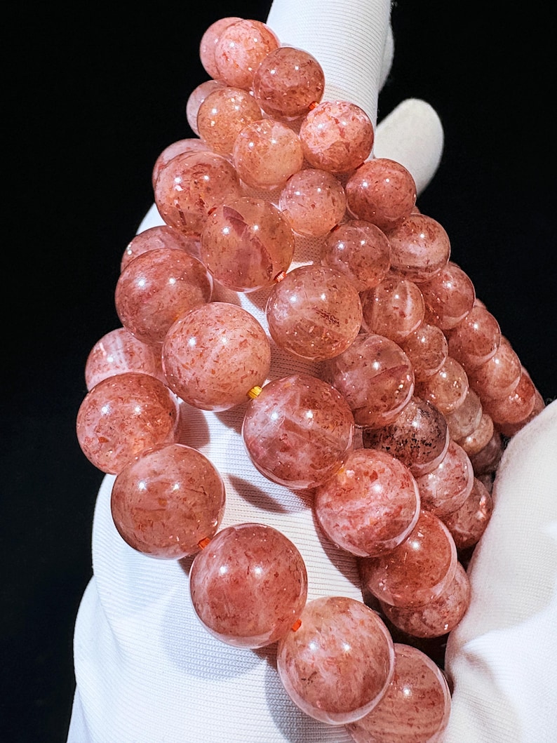 May include: A close-up of a bracelet made of round, reddish-orange beads. The beads have a speckled appearance and are strung together on a thin, orange cord. The bracelet is held against a white background.