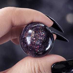 Puede incluir: Una piedra esférica pulida que presenta un color base morado oscuro y negro con motas iridiscentes y multicolores similares a la purpurina que captan la luz. La superficie es lisa y reflectante, mostrando una densa distribución de inclusiones brillantes.