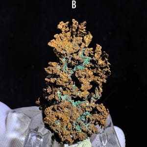 Puede incluir: Primer plano de un esp&eacute;cimen de mineral de cobre con una superficie rugosa y texturizada. El mineral muestra una mezcla de tonos marrones y verdes, con la letra "B" en blanco en la parte superior. El esp&eacute;cimen est&aacute; montado sobre una base clara e inclinada.