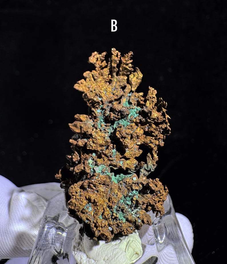 Puede incluir: Primer plano de un esp&eacute;cimen de mineral de cobre con una superficie rugosa y texturizada. El mineral muestra una mezcla de tonos marrones y verdes, con la letra "B" en blanco en la parte superior. El esp&eacute;cimen est&aacute; montado sobre una base clara e inclinada.