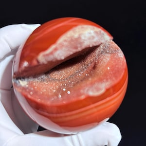 Op de afbeelding: Een rode agaat geode bol met een natuurlijke opening die het kristallijne interieur onthult. De bol heeft een gladde, gepolijste buitenkant en een ruwe, getextureerde binnenkant.