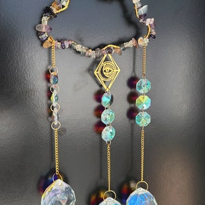 May include: A gold-toned crystal suncatcher with a cloud-shaped frame made of iridescent fluorite stones. The suncatcher has three strands of crystals, each ending in a large iridescent crystal ball.