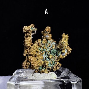 Puede incluir: Primer plano de un esp&eacute;cimen mineral de color dorado con parches verdes y azules. El mineral est&aacute; montado sobre una base rectangular transparente. La letra "A" es visible en la parte superior.