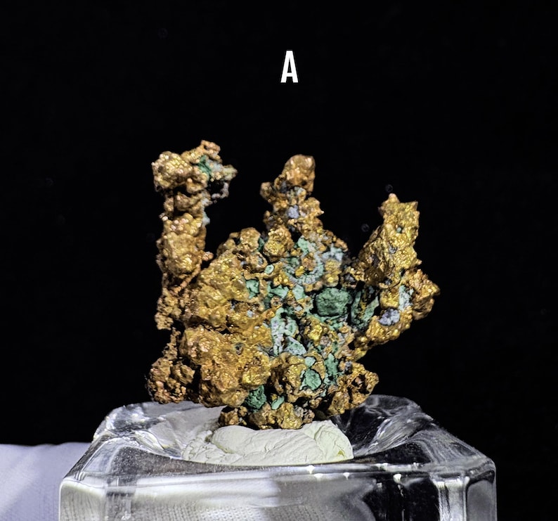 Puede incluir: Primer plano de un esp&eacute;cimen mineral de color dorado con parches verdes y azules. El mineral est&aacute; montado sobre una base rectangular transparente. La letra "A" es visible en la parte superior.