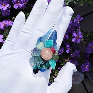 May include: A ring featuring a cluster of assorted raw and polished gemstones in shades of blue, green, and teal, centred around a smooth, round, pale peach-coloured stone, all set against a background of small purple flowers.