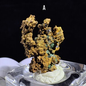 Puede incluir: Primer plano de un esp&eacute;cimen mineral de color dorado con una superficie rugosa y texturizada. El mineral tiene un grupo de formas irregulares con toques de verde y est&aacute; montado sobre una base blanca dentro de un soporte de vidrio transparente. La letra "A" es visible en la parte superior central.