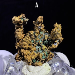Puede incluir: Primer plano de un esp&eacute;cimen mineral de color dorado con acentos verdes y azules. El mineral tiene una superficie texturizada e irregular y est&aacute; montado sobre una base blanca. La letra "A" es visible en la parte superior central.