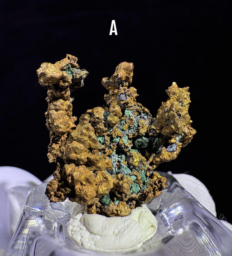 Puede incluir: Primer plano de un esp&eacute;cimen mineral de color dorado con acentos verdes y azules. El mineral tiene una superficie texturizada e irregular y est&aacute; montado sobre una base blanca. La letra "A" es visible en la parte superior central.